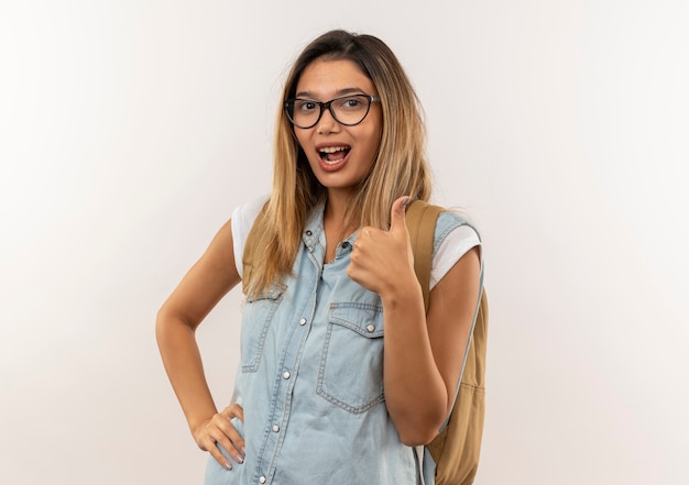 Foto gratuita fiduciosa giovane bella studentessa con gli occhiali e borsa posteriore mettendo la mano sulla vita e mostrando il pollice in alto isolato su bianco con spazio di copia