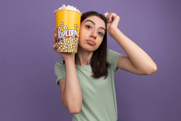 Уверенная молодая красивая женщина, держащая ведро попкорна и кусок попкорна, касаясь головы ведром попкорна и рукой, смотрящей вперед, изолированной на фиолетовой стене с копией пространства