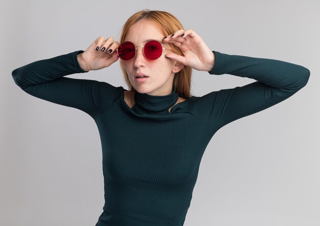 Confident young redhead ginger girl with freckles in sun glasses holding hands close to face isolated on white wall with copy space