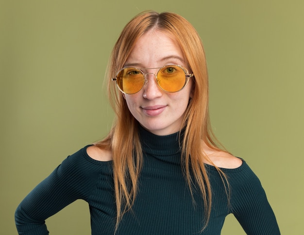 Confident young redhead ginger girl with freckles in sun glasses looking at camera on olive green