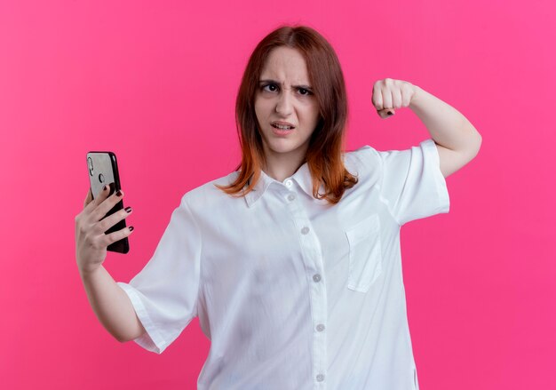 Confident young redhead girl holding phone and showing strong gesture isolated on pink wall