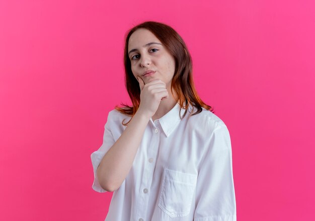 Confident young redhead girl putting hand under chin isolated on pink background