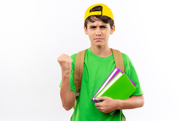 Confident young school boy wearing backpack with cap holding books showing yes gesture 