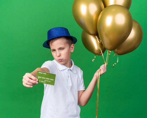confident young slavic boy with blue party hat holding helium balloons and credit card isolated on green wall with copy space