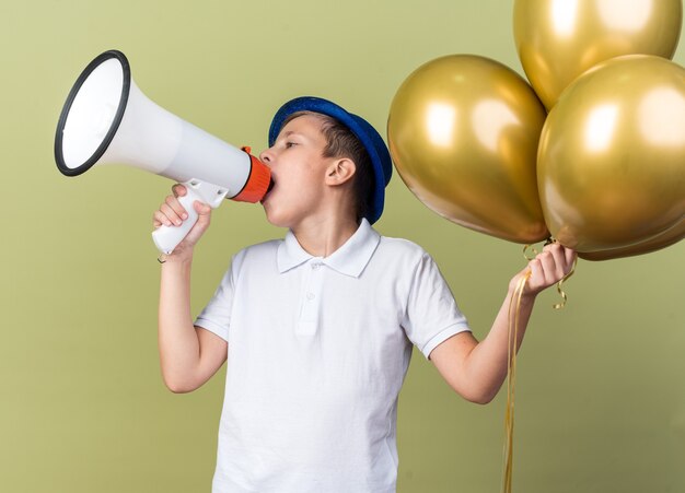 confident young slavic boy with blue party hat holding helium balloons and speaking into loud speaker looking at side isolated on olive green wall with copy space