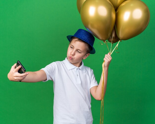 confident young slavic boy with blue party hat holding helium balloons taking selfie on phone isolated on green wall with copy space