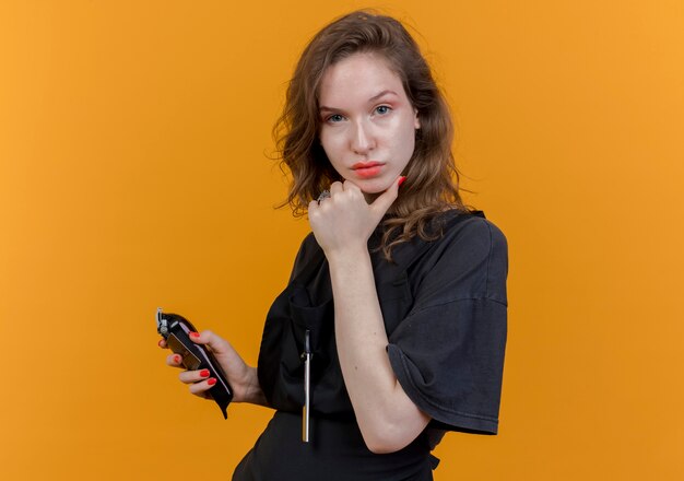 Confident young slavic female barber wearing uniform holding hair clippers and credit card touching chin