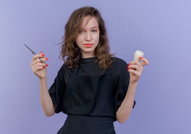 Confident young slavic female barber wearing uniform holding shaving brush and scissors isolated on purple background with copy space