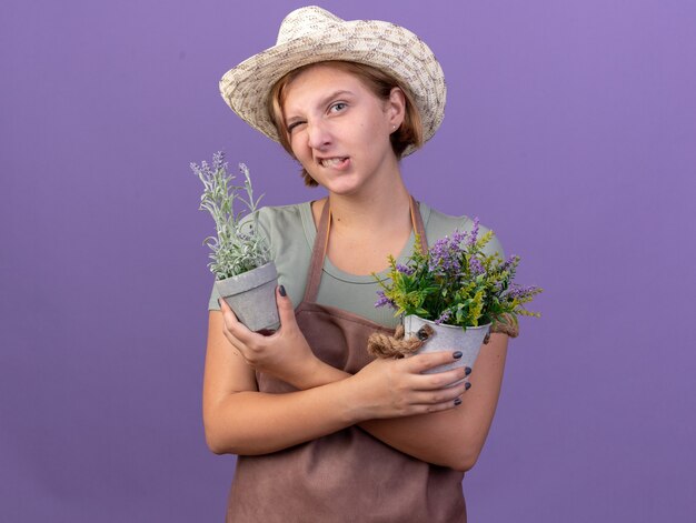 Confident young slavic female gardener wearing gardening hat blinks eye and holds flowers in flowerpots on purple