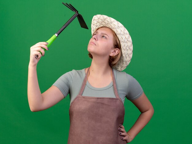 Confident young slavic female gardener wearing gardening hat holding and looking at hoe rake on green