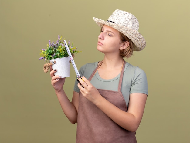 Confident young slavic female gardener wearing gardening hat measuring flowerpot with tape measure on olive green