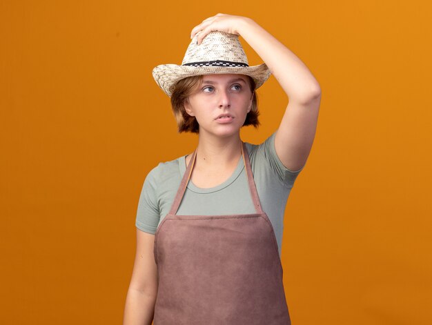 원예 모자를 쓰고 자신감이 젊은 슬라브 여성 정원사는 오렌지 측면을보고 모자에 손을 넣습니다.