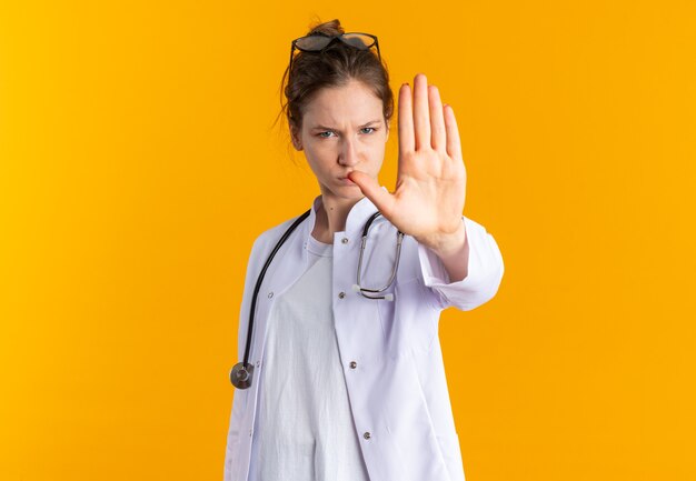 Confident young slavic girl in doctor uniform with stethoscope gesturing stop hand sign isolated on orange wall with copy space