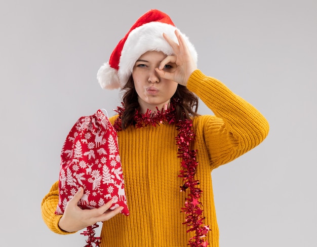 Confident young slavic girl with santa hat and with garland around neck holding christmas gift bag and looking at camera through fingers isolated on white background with copy space