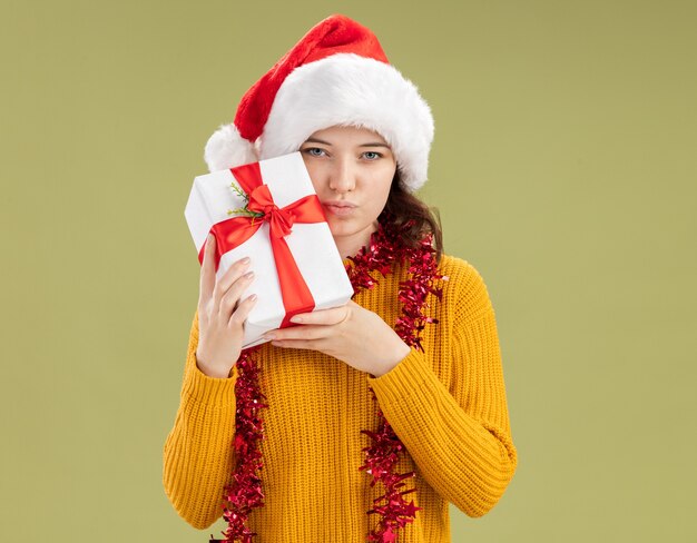 Confident young slavic girl with santa hat and with garland around neck holding christmas gift box isolated on olive green wall with copy space