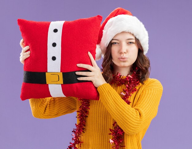 Confident young slavic girl with santa hat and with garland around neck holding decorated pillow isolated on purple background with copy space