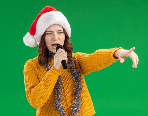 confident young slavic girl with santa hat and with garland around neck holding mic and pointing at side isolated on green background with copy space