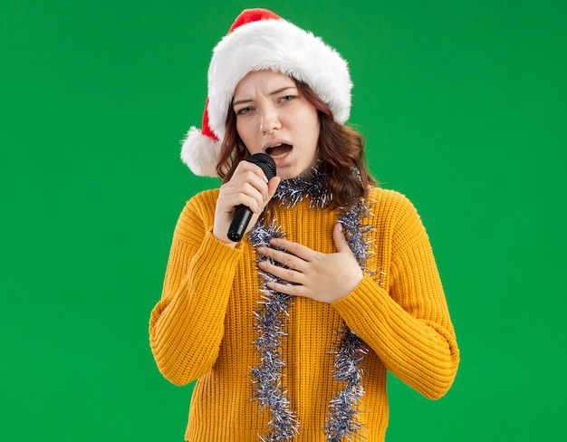 Free photo confident young slavic girl with santa hat and with garland around neck holds mic pretending to sing isolated on green background with copy space