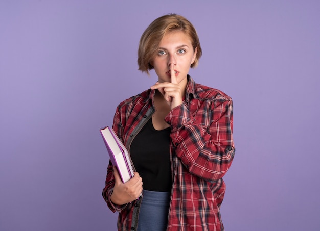 Confident young slavic student girl holds book and notebook gestures silence sign solated on purple background with copy space
