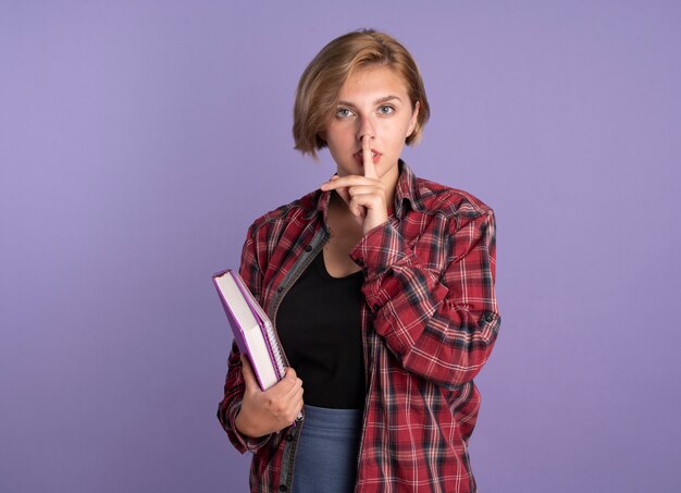 Confident young slavic student girl holds book and notebook gestures silence sign solated on purple background with copy space