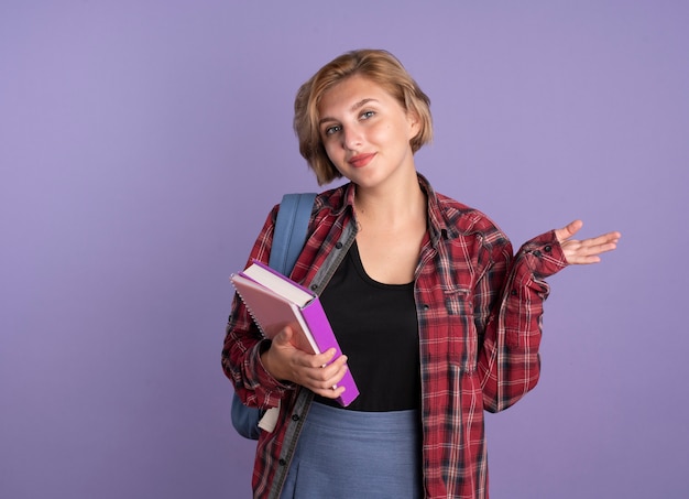 Confident young slavic student girl wearing backpack holds book and notebook keeps hand open 