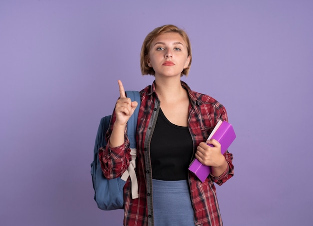 Confident young slavic student girl wearing backpack holds book and notebook points up 