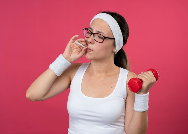 Confident young sporty girl in optical glasses wearing headband and wristbands holds dumbbell and pretends to smoke cigarette 
