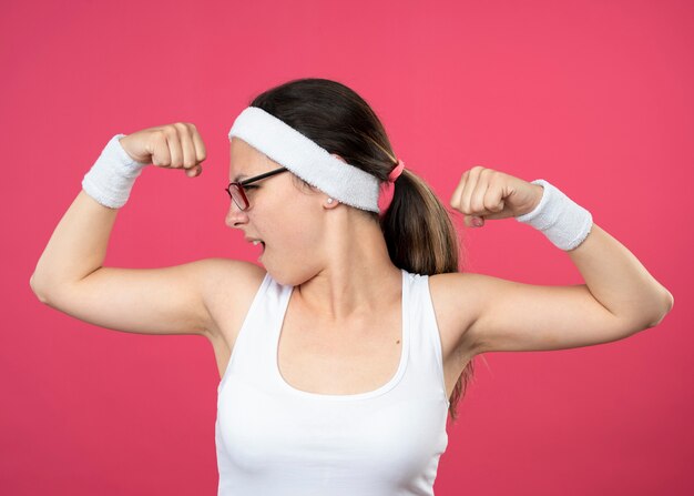 Confident young sporty girl in optical glasses wearing headband and wristbands tenses biceps and looks at side 