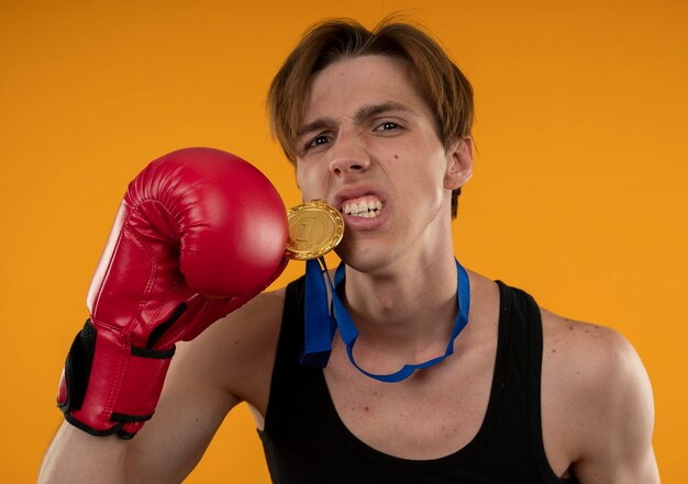 Confident young sporty guy wearing boxing gloves and holding medal isolated on orange wall