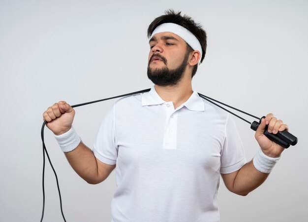 Confident young sporty man looking at side wearing headband and wristband holding jumb rope on behind neck isolated on white wall