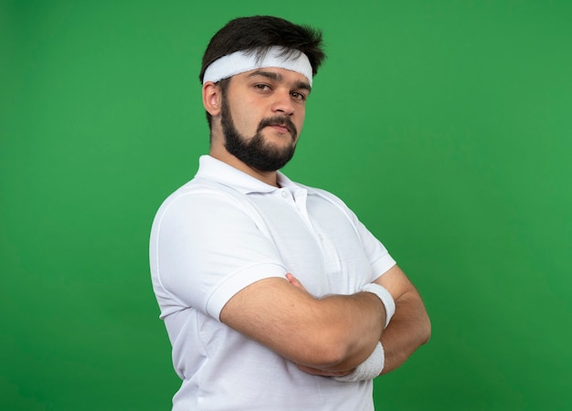 Confident young sporty man wearing headband and wristband crossing hands isolated on green wall