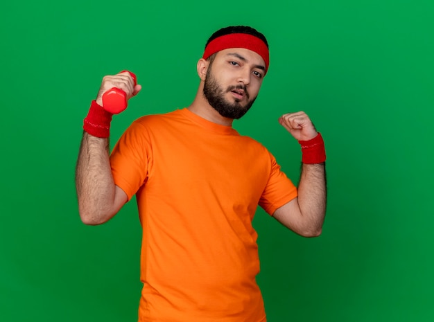 Confident young sporty man wearing headband and wristband exercising with dumbbell showing strong gesture isolated on green background