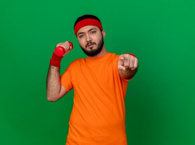 Confident young sporty man wearing headband and wristband exercising with dumbbell showing you gesture isolated on green background