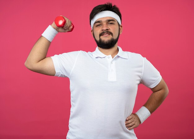 Confident young sporty man wearing headband and wristband holding dumbbell and putting hand on hip isolated on pink