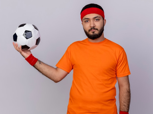 Free photo confident young sporty man wearing headband and wristband holding out ball at side isolated on white background