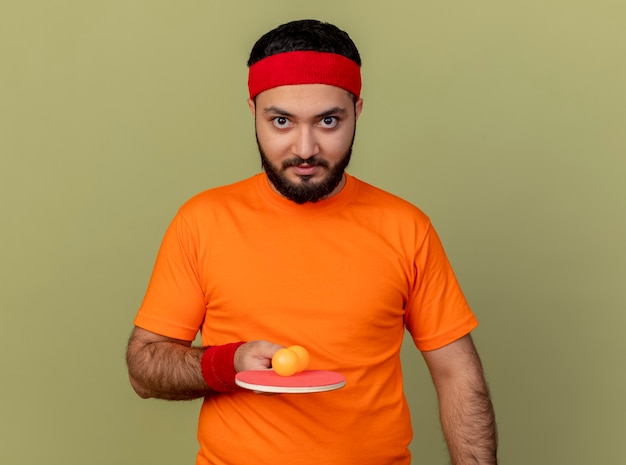 Confident young sporty man wearing headband and wristband holding ping pong racket with ball isolated on olive green background