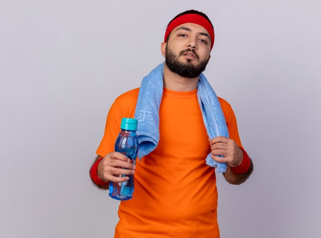 Confident young sporty man wearing headband and wristband holding water bottle with towel on shoulder isolated on white background