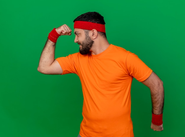 Foto gratuita fiducioso giovane uomo sportivo che indossa la fascia e il braccialetto che mostra un forte gesto isolato su sfondo verde