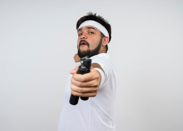 Confident young sporty man wearing headband and wristband stretching a jump rope isolated on white with copy space