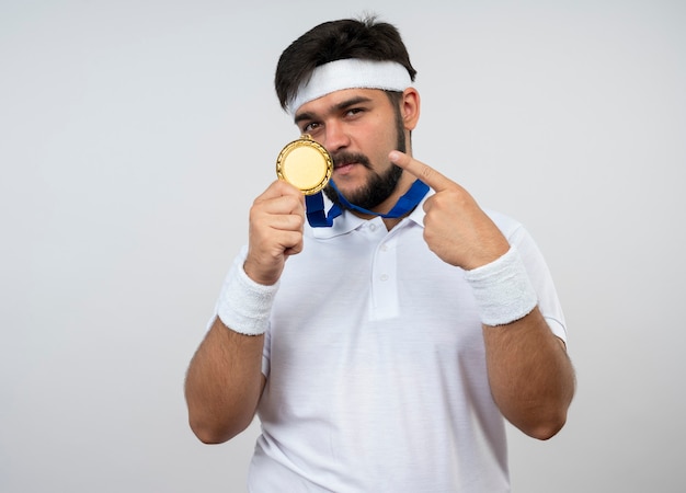 Confident young sporty man wearing headband and wristband wearing and points at medal isolated on white wall