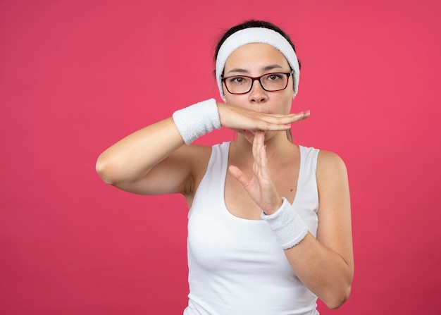 Уверенная молодая спортивная женщина в оптических очках, носящая повязку на голову и браслеты, жестикулирует по тайм-ауту, изолированному на розовой стене