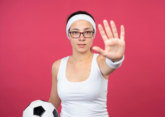 Уверенная молодая спортивная женщина в оптических очках с повязкой на голову и браслетами держит мяч и жестами останавливает знак рукой, изолированную на розовой стене