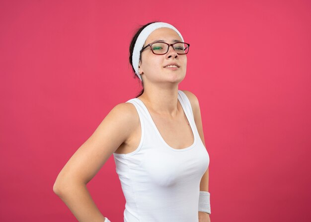 Confident young sporty woman in optical glasses wearing headband and wristbands puts hands on waist and looks at front isolated on pink wall