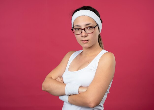 Confident young sporty woman in optical glasses wearing headband and wristbands stands with crossed arms isolated on pink wall