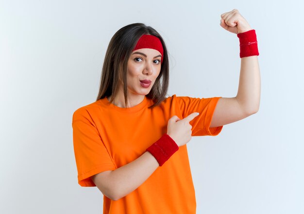 Confident young sporty woman wearing headband and wristbands  doing strong gesture pointing at her muscles isolated on white wall with copy space