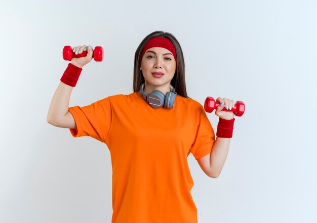 Confident young sporty woman wearing headband and wristbands and headphones on neck  raising up dumbbells isolated on white wall with copy space