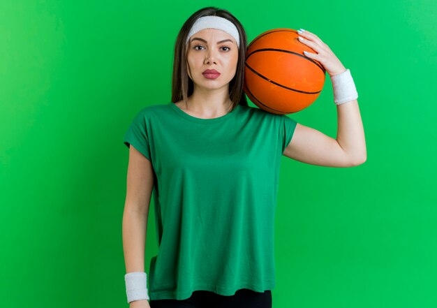 Confident young sporty woman wearing headband and wristbands holding basketball ball on shoulder looking 