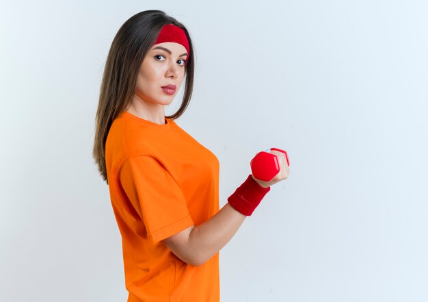 Confident young sporty woman wearing headband and wristbands standing in profile view holding dumbbell looking isolated