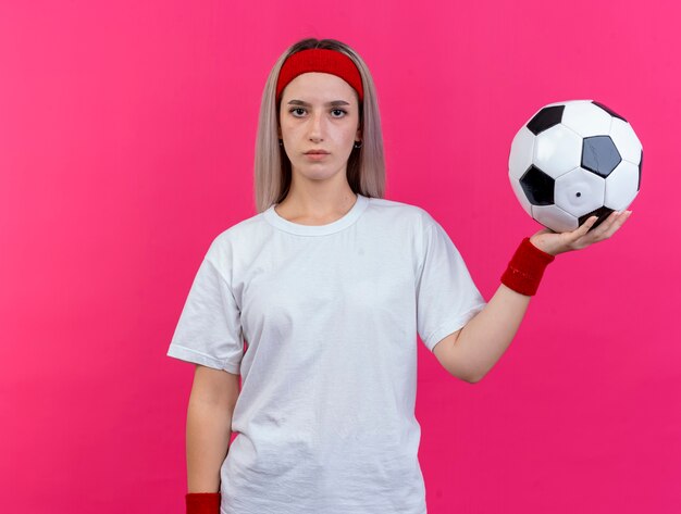 머리띠와 팔찌를 착용하는 중괄호와 자신감이 젊은 스포티 한 여자는 분홍색 벽에 고립 된 공을 보유