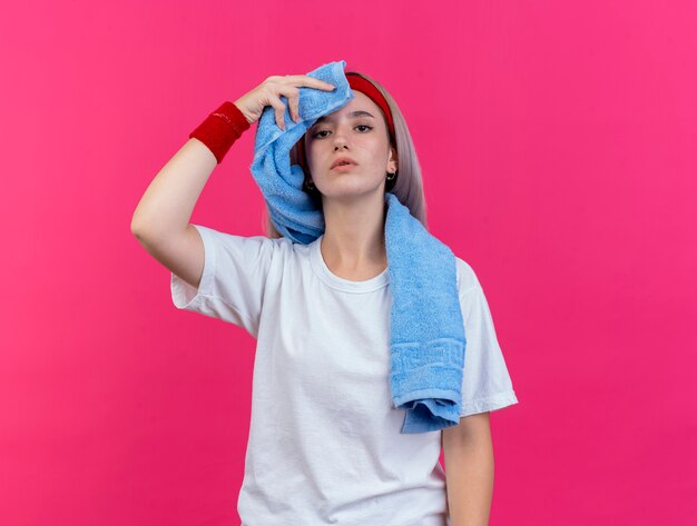 Confident young sporty woman with braces wearing headband and wristbands holds and wipes forehead with towel isolated on pink wall